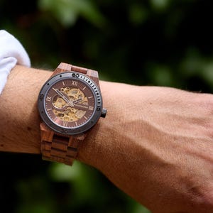 May include: A wooden wristwatch with a brown face and band. The watch has a visible mechanical movement and Roman numeral hour markers. The watch is on a wrist, with a white shirt sleeve visible in the background.