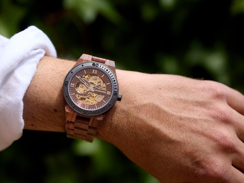 May include: A wooden wristwatch with a brown face and band. The watch has a visible mechanical movement and Roman numeral hour markers. The watch is on a wrist, with a white shirt sleeve visible in the background.