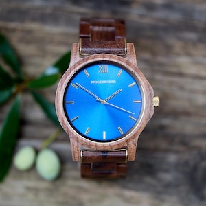 May include: A wooden wristwatch with a vibrant blue face and gold-colored hands and accents. The watch features the word "WOODPECKER" on the face and a wooden band. The watch is displayed on a wooden surface.