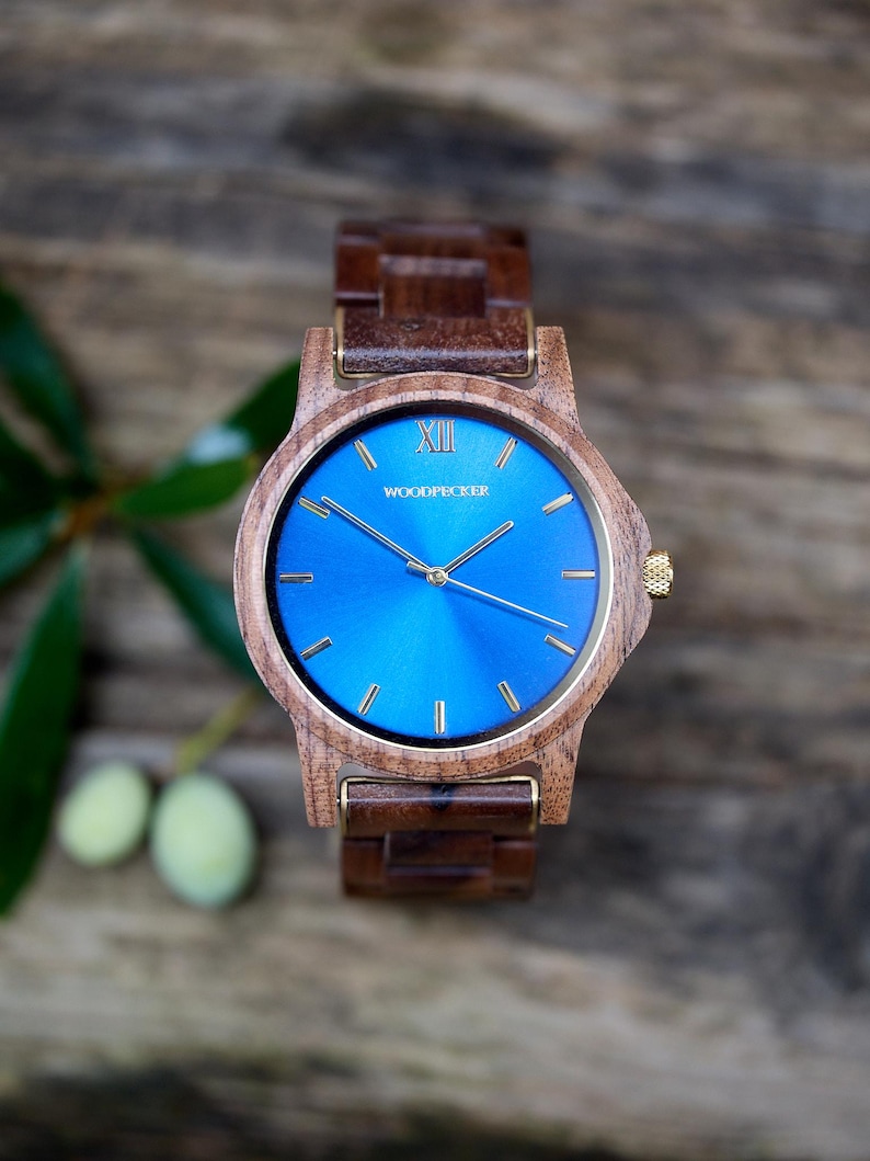 May include: A wooden wristwatch with a vibrant blue face and gold-colored hands and accents. The watch features the word "WOODPECKER" on the face and a wooden band. The watch is displayed on a wooden surface.