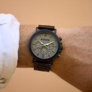 Puede incluir: Un reloj de pulsera cron&oacute;grafo negro con una esfera de madera y una correa de metal negro. La esfera del reloj tiene marcadores de hora en n&uacute;meros romanos y una ventana de fecha.