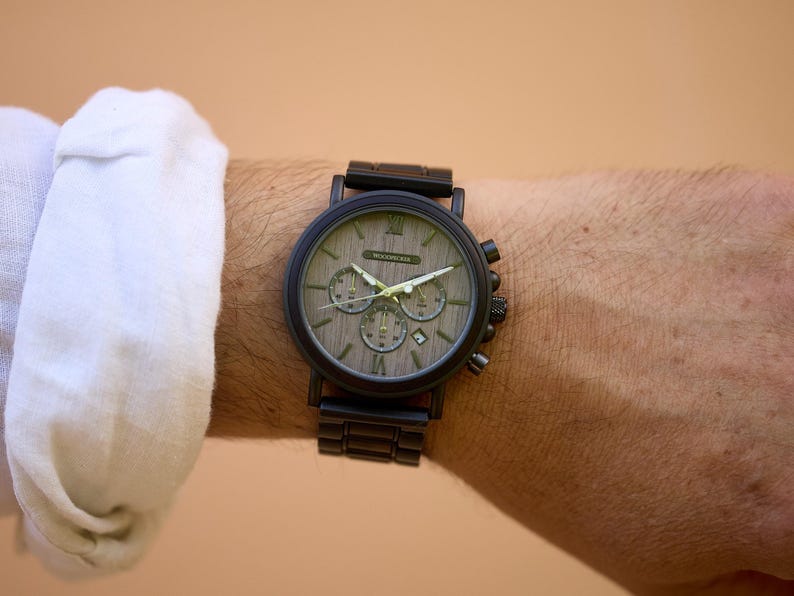 Puede incluir: Un reloj de pulsera cron&oacute;grafo negro con una esfera de madera y una correa de metal negro. La esfera del reloj tiene marcadores de hora en n&uacute;meros romanos y una ventana de fecha.