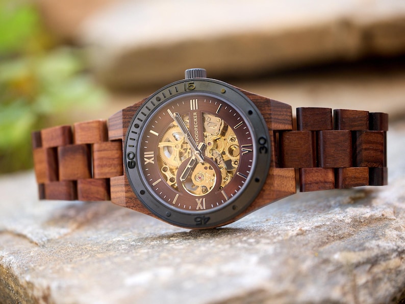 May include: A wooden wristwatch with a dark brown face and a visible mechanical movement. The watch has a dark gray bezel and a wooden band. The watch is displayed on a stone surface.
