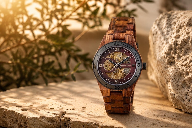 May include: A wooden wristwatch with a brown band and a burgundy face. The watch face has visible gears and Roman numerals. The watch is displayed on a textured stone surface with foliage in the background.