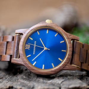 May include: A wooden wristwatch with a blue face and gold-colored hands and accents. The watch has a brown wooden band and the word "WOODPECKER" is printed on the watch face. The watch is displayed on a wooden surface.