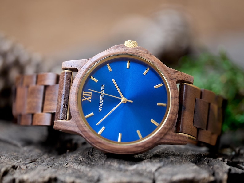 May include: A wooden wristwatch with a blue face and gold-colored hands and accents. The watch has a brown wooden band and the word "WOODPECKER" is printed on the watch face. The watch is displayed on a wooden surface.