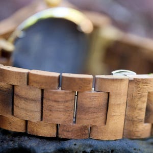 May include: Close-up of a wooden watch with a natural wood grain finish. The watch band is made of interlocking wooden blocks, and the watch face is partially visible. The watch has a silver clasp.