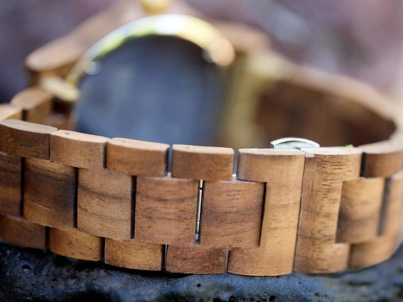 May include: Close-up of a wooden watch with a natural wood grain finish. The watch band is made of interlocking wooden blocks, and the watch face is partially visible. The watch has a silver clasp.