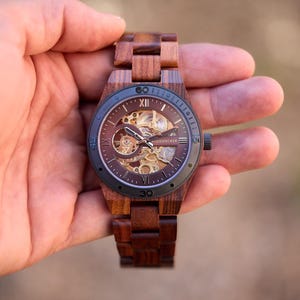 Puede incluir: Un reloj de pulsera de madera marr&oacute;n con una esfera negra y un movimiento de esqueleto. El reloj tiene marcadores de hora en n&uacute;meros romanos y una correa de cuero negra.