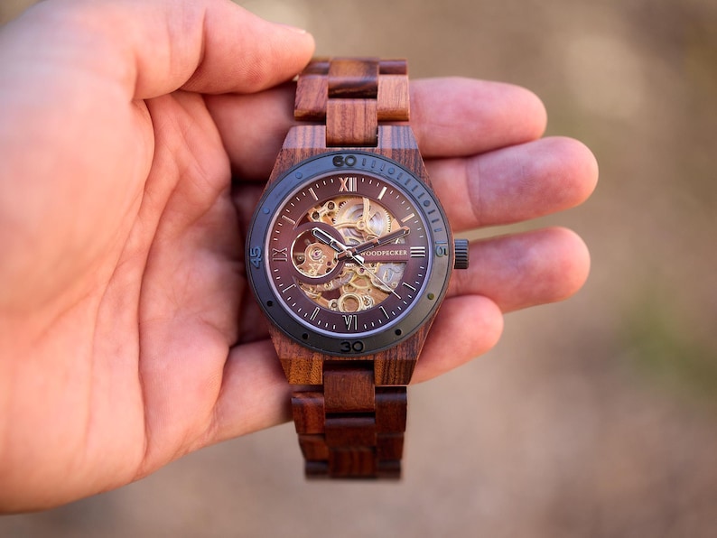 Puede incluir: Un reloj de pulsera de madera marr&oacute;n con una esfera negra y un movimiento de esqueleto. El reloj tiene marcadores de hora en n&uacute;meros romanos y una correa de cuero negra.