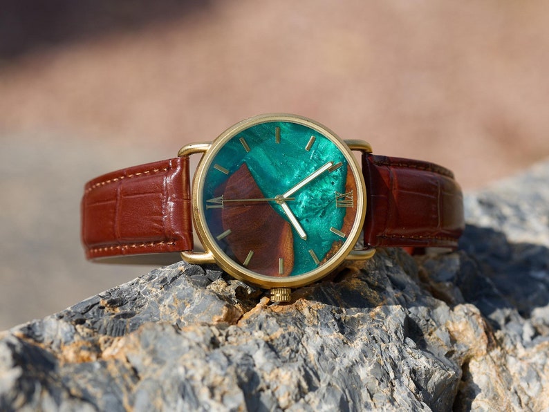 Puede incluir: Un reloj de pulsera dorado con una correa de cuero marr&oacute;n. La esfera del reloj presenta un dise&ntilde;o &uacute;nico con tonos verdes y marrones. Los n&uacute;meros romanos marcan las horas. El reloj se muestra sobre una superficie rocosa texturizada.