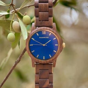 May include: A wooden wristwatch with a blue face and gold accents. The watch has a dark brown wooden band and a round face with gold hands and markers. The watch face has the text "XII" and "WOODPECKER". The watch is displayed against a blurred green background.