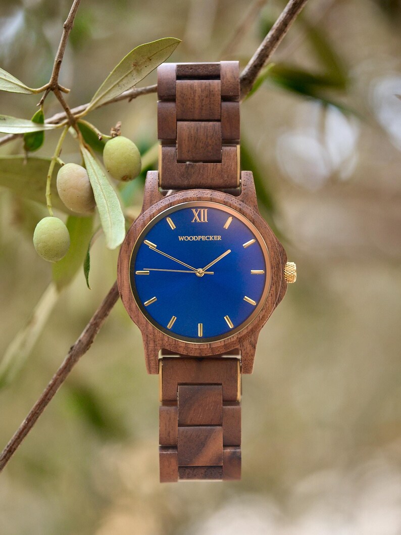 May include: A wooden wristwatch with a blue face and gold accents. The watch has a dark brown wooden band and a round face with gold hands and markers. The watch face has the text "XII" and "WOODPECKER". The watch is displayed against a blurred green background.