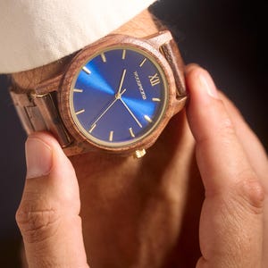 May include: A wooden wristwatch with a blue face and gold accents. The watch has a brown wooden band and a gold crown. The watch face has gold hands and markers. The watch is being held by a person.