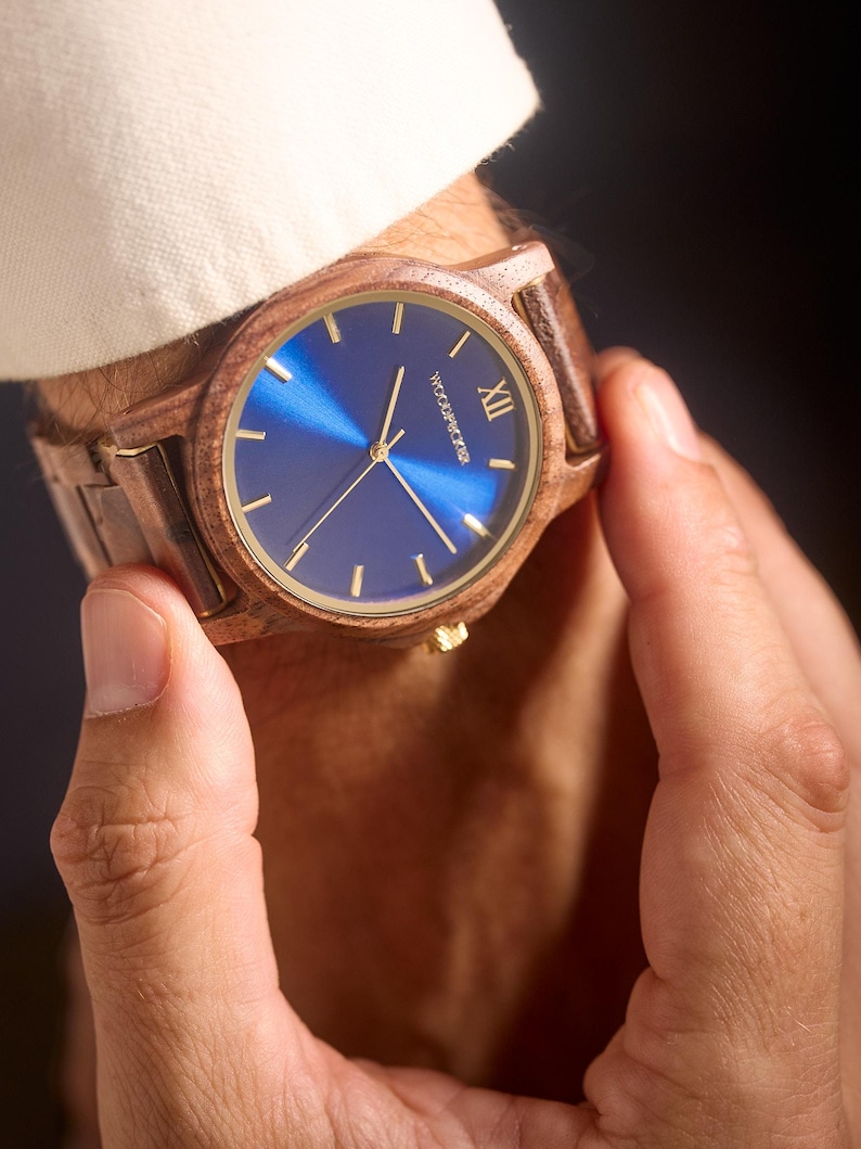 May include: A wooden wristwatch with a blue face and gold accents. The watch has a brown wooden band and a gold crown. The watch face has gold hands and markers. The watch is being held by a person.