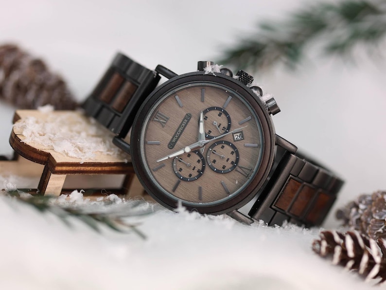 Pu&ograve; includere: Un orologio cronografo in legno marrone con cinturino in metallo nero. Il quadrante dell'orologio ha indici orari in numeri romani e una finestra della data alle 4.