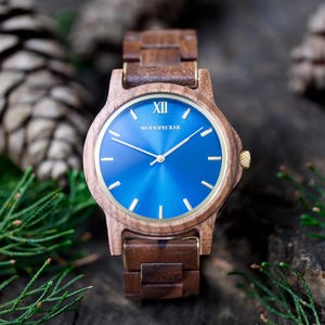 May include: A wooden wristwatch with a blue face and gold accents. The watch has a brown wooden band and a gold crown. The watch face has gold hands and markers. The word "WOODPECKER" is printed on the watch face.