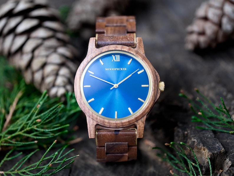 May include: A wooden wristwatch with a blue face and gold accents. The watch has a brown wooden band and a gold crown. The watch face has gold hands and markers. The word "WOODPECKER" is printed on the watch face.