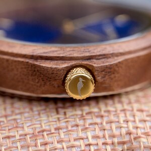 May include: Close-up of a wooden watch with a blue face. The watch has a gold-colored crown with a seahorse engraving. The watch is resting on a woven surface.