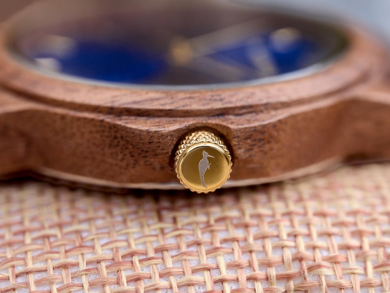 May include: Close-up of a wooden watch with a blue face. The watch has a gold-colored crown with a seahorse engraving. The watch is resting on a woven surface.