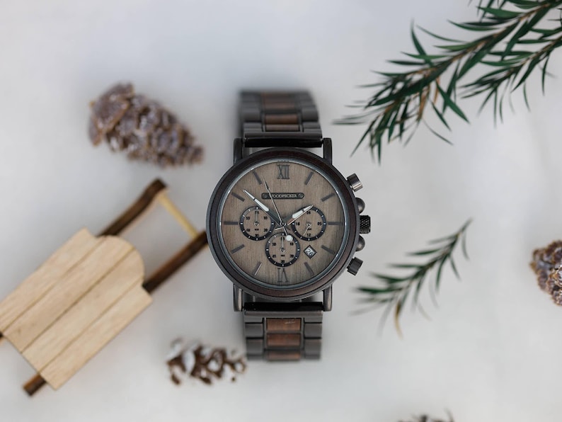 Pu&ograve; includere: Un orologio da polso in legno marrone con quadrante cronografo e cinturino in legno marrone. L'orologio ha indici orari in numeri romani e una finestrella per la data alle 4.