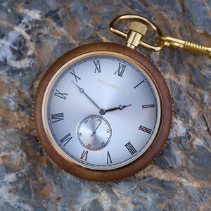 May include: A gold pocket watch with Roman numeral markings on a white face. The watch has a brown wooden case and a gold chain.