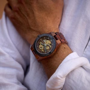 Puede incluir: Un reloj de pulsera de madera marr&oacute;n con una esfera negra y un movimiento autom&aacute;tico visible. El reloj tiene una correa de cuero marr&oacute;n.