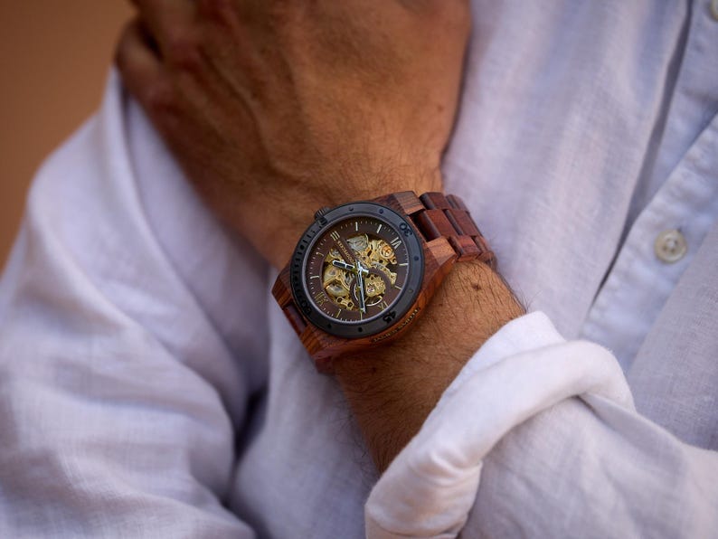 Puede incluir: Un reloj de pulsera de madera marr&oacute;n con una esfera negra y un movimiento autom&aacute;tico visible. El reloj tiene una correa de cuero marr&oacute;n.