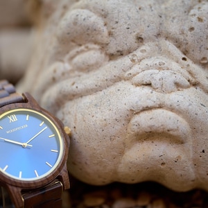 May include: A wooden wristwatch with a blue face and gold accents. The watch has a brown wooden band and a gold-colored bezel. The watch face displays the brand name "Woodpecker". The watch is next to a stone sculpture.