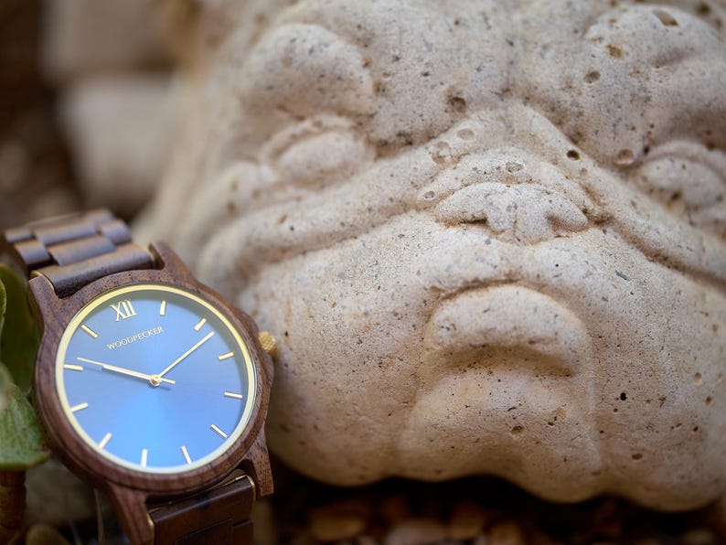 May include: A wooden wristwatch with a blue face and gold accents. The watch has a brown wooden band and a gold-colored bezel. The watch face displays the brand name "Woodpecker". The watch is next to a stone sculpture.