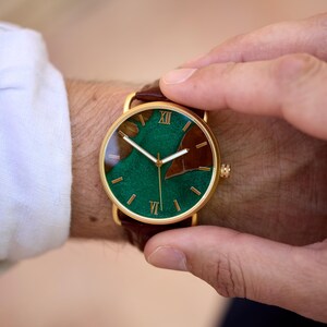 Puede incluir: Un reloj dorado con esfera verde y marr&oacute;n y correa de cuero marr&oacute;n. La esfera del reloj presenta n&uacute;meros romanos y un dise&ntilde;o &uacute;nico. El reloj se lleva en la mu&ntilde;eca.