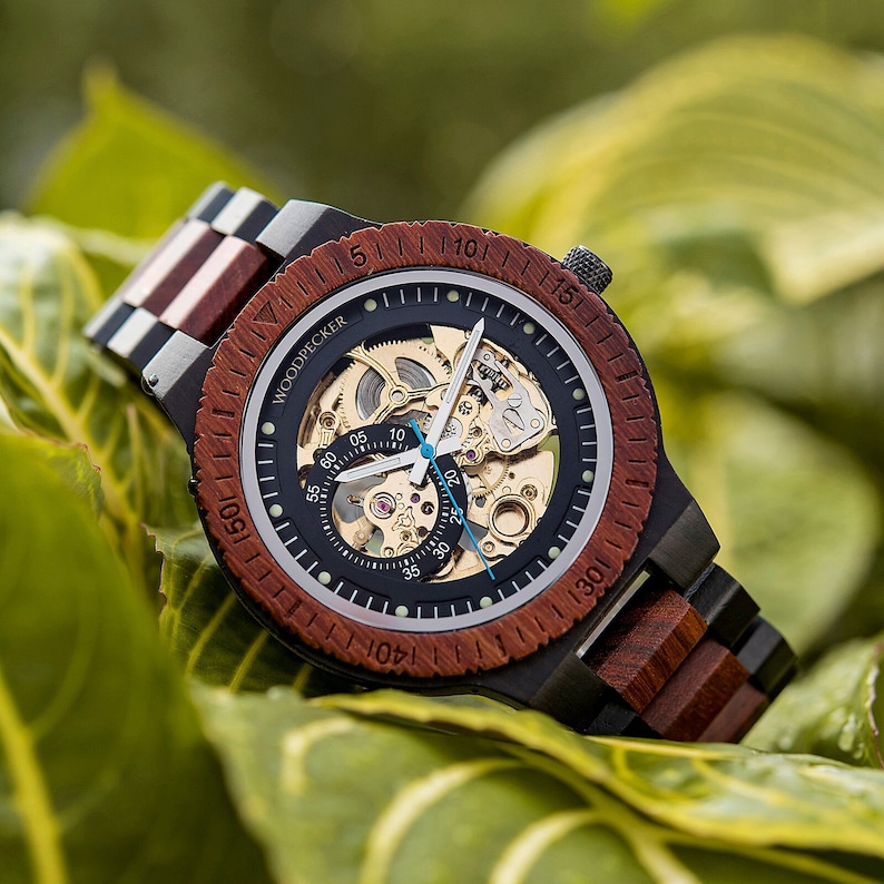 May include: A black and brown wooden wristwatch with a skeleton dial and gold-toned hands. The watch face has a white background with black markings and the text "WOODPECKER" printed on it.
