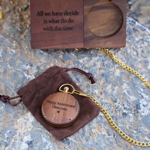 May include: A wooden pocket watch with a gold chain and a brown velvet pouch. The watch face is engraved with the words "Happy Anniversary I love you".