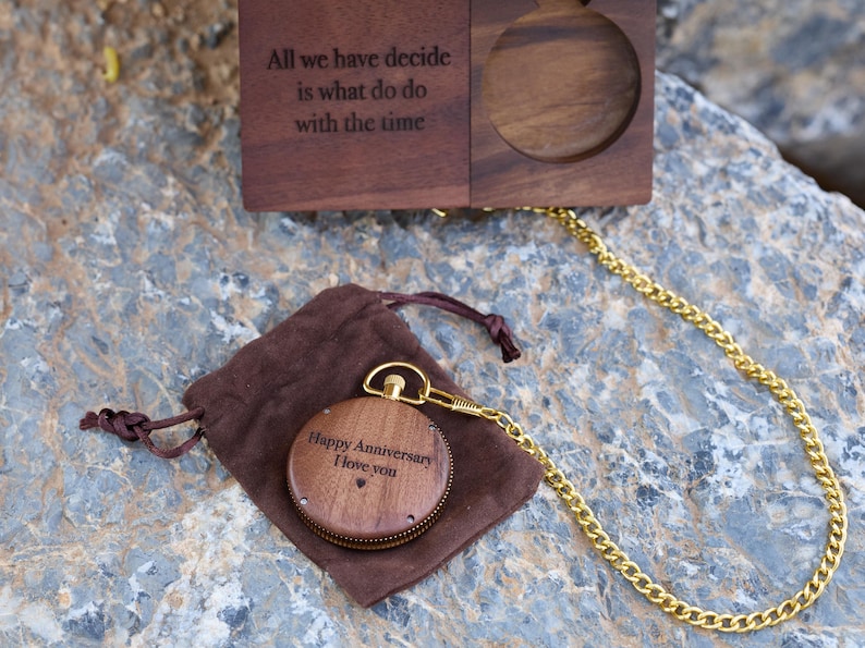 May include: A wooden pocket watch with a gold chain and a brown velvet pouch. The watch face is engraved with the words "Happy Anniversary I love you".