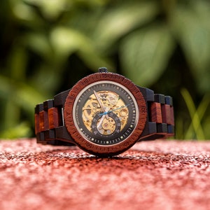 May include: A wooden wristwatch with a black and red wood band. The watch face is a skeleton design with a visible mechanical movement. The watch face has a black background with gold accents and white markings. The watch face also has a blue second hand.