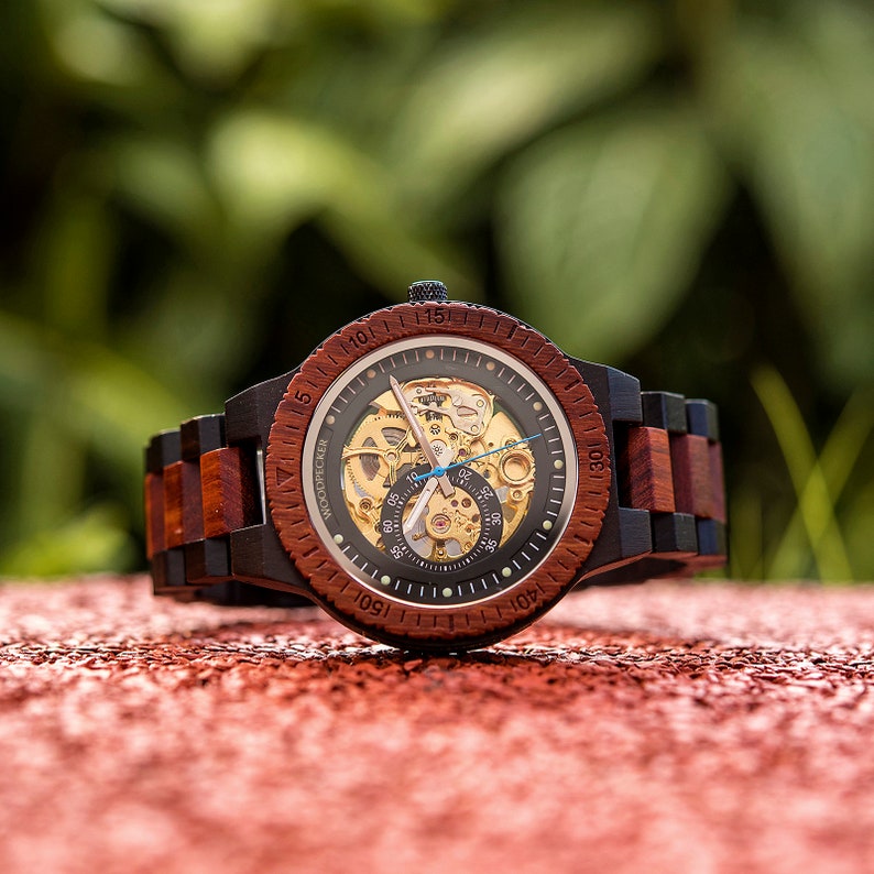 May include: A wooden wristwatch with a black and red wood band. The watch face is a skeleton design with a visible mechanical movement. The watch face has a black background with gold accents and white markings. The watch face also has a blue second hand.