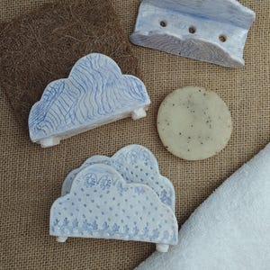 Peut inclure: Ensemble de porte-savons en céramique faits à la main et une barre de savon. Les porte-savons en forme de nuage sont peints en bleu et blanc, avec des motifs décoratifs. La barre de savon est ronde et de couleur claire. Les articles sont disposés sur une surface texturée de tons neutres.