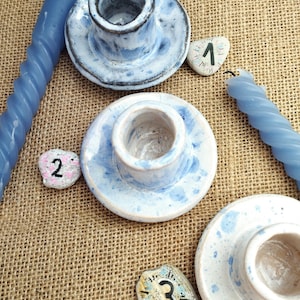 May include: Ceramic candle holders with blue and white speckled glaze. The image shows three candle holders, two blue spiral candles, and three numbered stones. The candle holders are on a burlap surface.