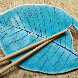 Peut inclure: Un plat de service en céramique turquoise en forme de feuille avec une surface texturée et des nervures bleu foncé. Deux baguettes en bois reposent sur le plat. Les baguettes ont des motifs floraux bleus aux extrémités. Le fond est en toile de jute beige.