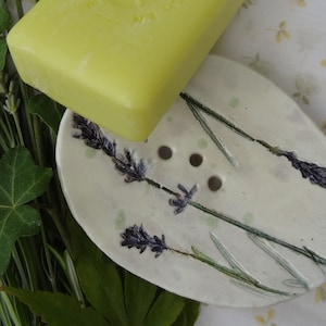 K&ouml;nnte beinhalten: Eine wei&szlig;e Keramik-Seifenschale mit einem Lavendel-Design. Die Schale hat vier Abflussl&ouml;cher und ist oval geformt. Eine gelbe Seifenstange liegt auf der Schale.