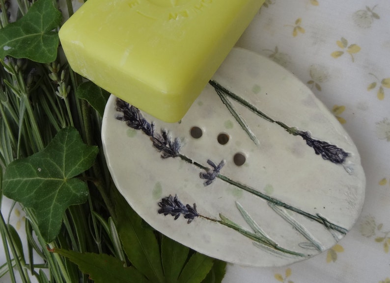 K&ouml;nnte beinhalten: Eine wei&szlig;e Keramik-Seifenschale mit einem Lavendel-Design. Die Schale hat vier Abflussl&ouml;cher und ist oval geformt. Eine gelbe Seifenstange liegt auf der Schale.
