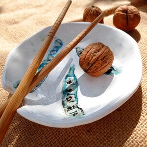 Peut inclure: Une assiette en céramique blanche avec des accents bleus et turquoise, accompagnée de baguettes en bois et d'une noix. L'assiette a une forme organique et convient pour servir des sushis ou des amuse-gueules. Les baguettes sont marron clair.