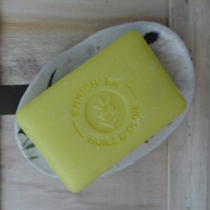 K&ouml;nnte beinhalten: Eine gelbe Seife mit dem Text "ENRICHI EN HUILE D'OLIVE" in einem Kreis auf der Vorderseite.