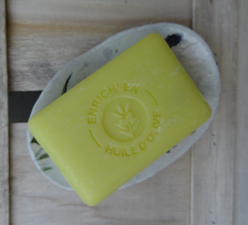 K&ouml;nnte beinhalten: Eine gelbe Seife mit dem Text "ENRICHI EN HUILE D'OLIVE" in einem Kreis auf der Vorderseite.