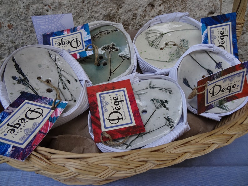 K&ouml;nnte beinhalten: Ein Weidenkorb gef&uuml;llt mit Keramik-Seifenschalen. Jede Schale zeigt ein anderes gepresstes Blumenmuster und ist in wei&szlig;es Seidenpapier eingewickelt. Die Schalen sind mit dem Text "atelier D&eacute;ge." beschriftet.