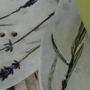 K&ouml;nnte beinhalten: Zwei wei&szlig;e Keramik-Seifenschalen mit einem floralen Design. Die Schalen haben einen erh&ouml;hten Rand und drei L&ouml;cher oben. Das Design zeigt gr&uuml;ne Bl&auml;tter und lila Lavendelbl&uuml;ten.
