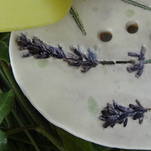 K&ouml;nnte beinhalten: Eine wei&szlig;e Keramik-Seifenschale mit einem Lavendel-Design. Die Schale hat drei Abflussl&ouml;cher und ist mit Lavendelbl&uuml;ten und Stielen verziert.