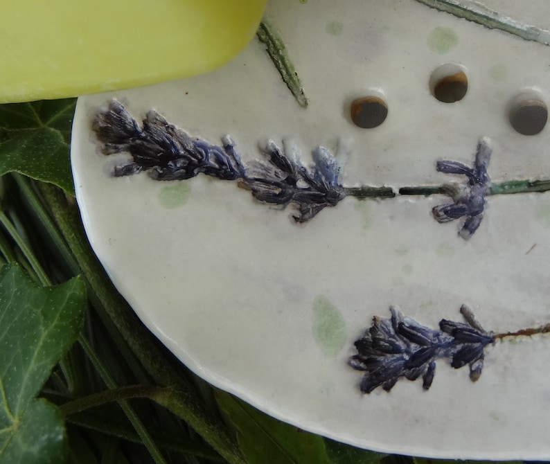 K&ouml;nnte beinhalten: Eine wei&szlig;e Keramik-Seifenschale mit einem Lavendel-Design. Die Schale hat drei Abflussl&ouml;cher und ist mit Lavendelbl&uuml;ten und Stielen verziert.