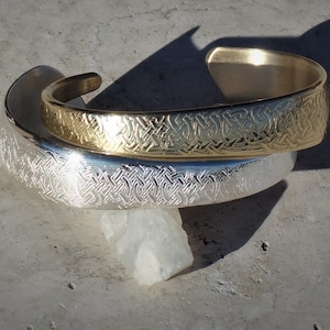 May include: Two silver and gold bracelets with an intricate Celtic knot design. The bracelets are stacked on top of each other.