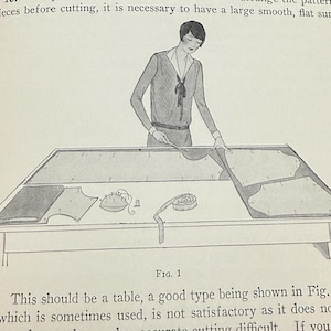 May include: An illustration from a vintage sewing guide shows a person arranging fabric on a large table. The table has a smooth surface, and various sewing tools are visible. The text reads "CUTTING" and "ARRANGING THE PATTERN".